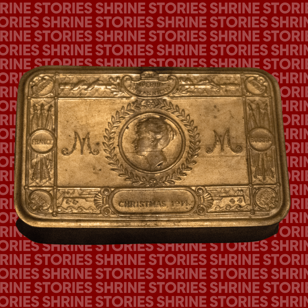 A brass tin with a face engraved. The face is surrounded by a wreath and on either side are the letters 'M'. 