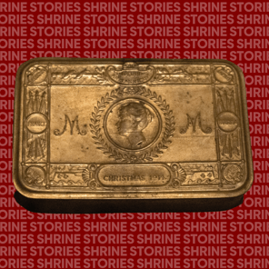 A brass tin with a face engraved. The face is surrounded by a wreath and on either side are the letters 'M'.