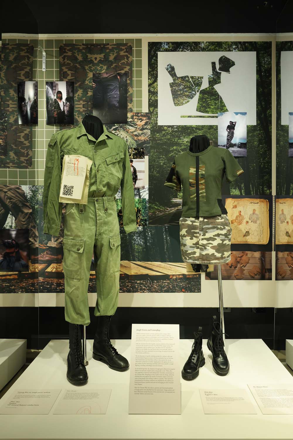 A display at the Shrine with two mannequins standing side by side. On the left there is a Vietnam era jungle greens uniform with a light green long-sleeved shirt and trousers. The mannequin is also wearing black lace up combat boots. Next to the jungle greens is a modern use of green and camouflage. The mannequin is wearing a green t-shirt with a camouflage rectangle in the centre and a camouflage mini skirt. There are black Doc Martin combat boots at the bottom. 