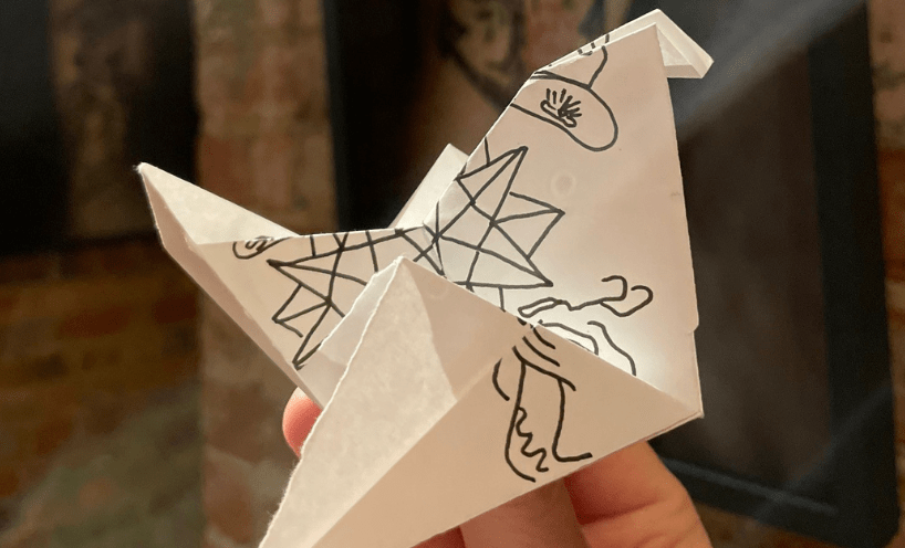 A white paper crane with tattoo-like designs drawn on it. In the background are brick pillars with images of tattoos on them. 