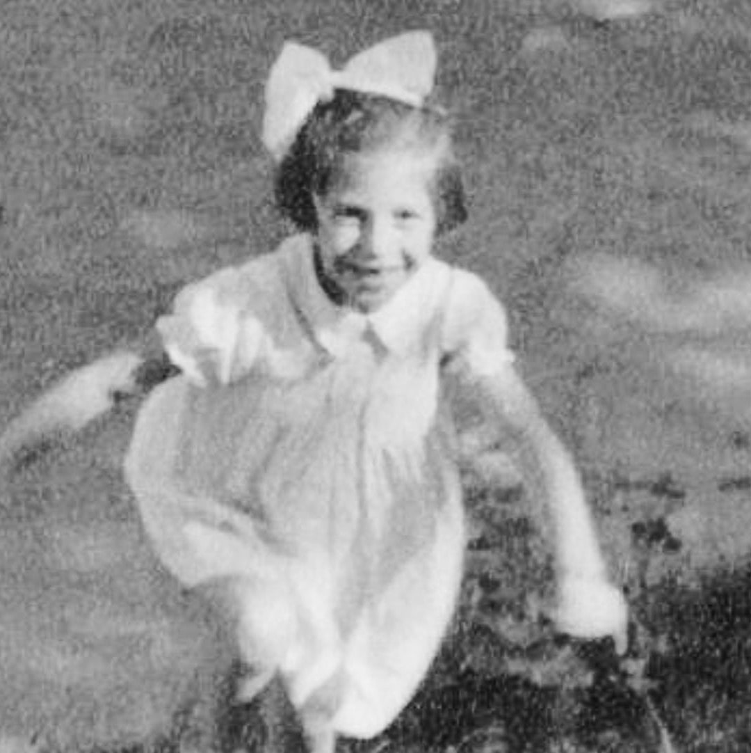 A black and white photo showing a young girl running in a field. She is wearing a white dress with a collar and a big white bow in her short hair. She is smiling as she moves. 