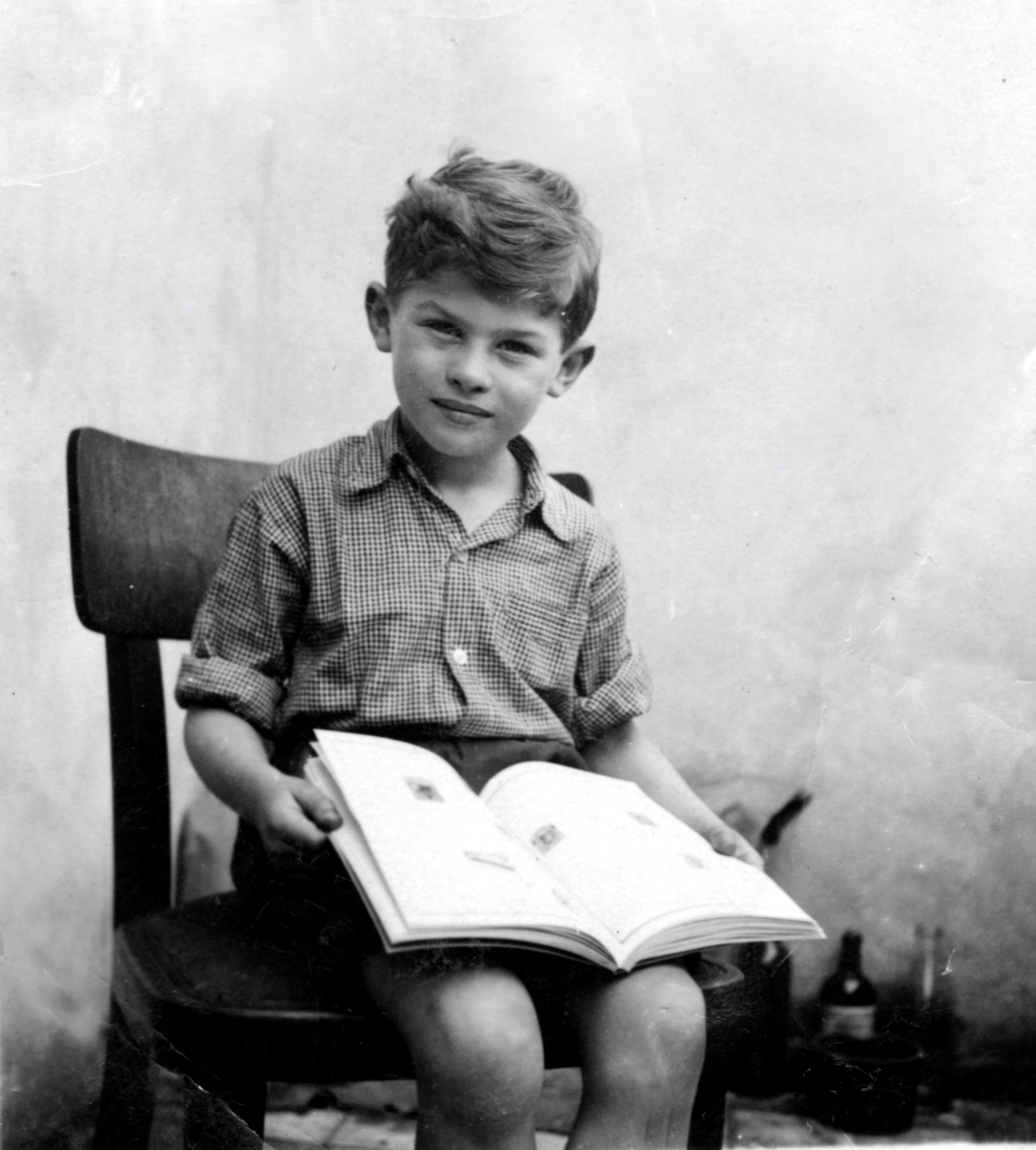 A black and white photograph of a young boy sitting on a chair. He is holding an open book and smiling into the camera. He is wearing a checked shirt with rolled up sleeves.