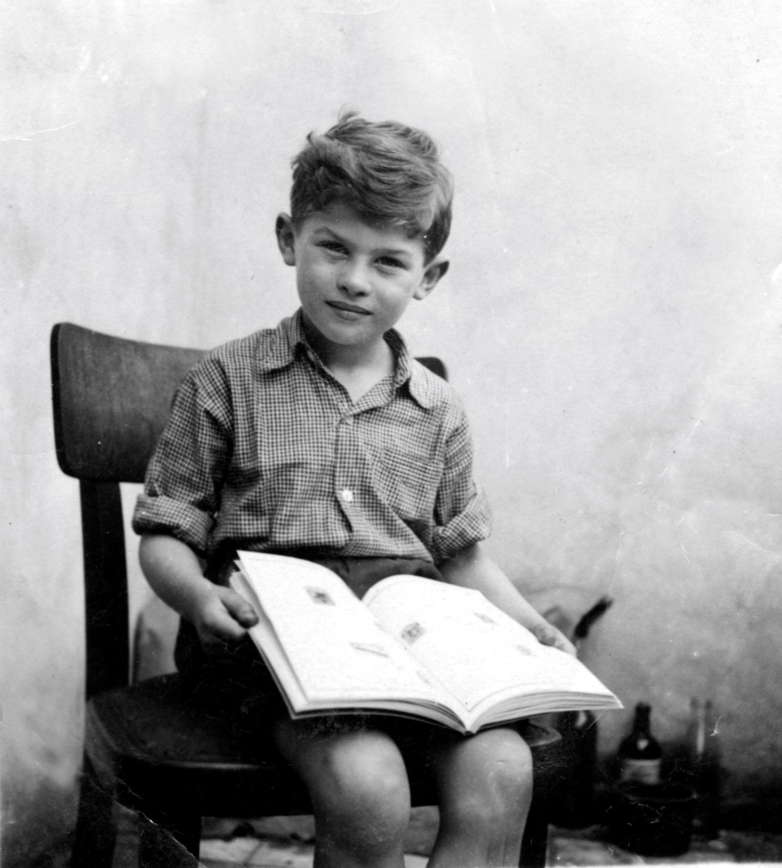 Garry Fabian A black and white photograph of a young boy sitting on a chair. He is holding an open book and smiling into the camera. He is wearing a checked shirt with rolled up sleeves.