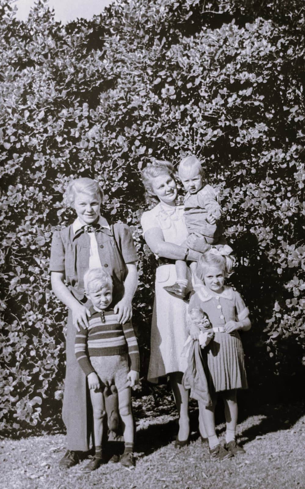 A group of five people standing together against a tall bush. On the left, there is a wman holding the shoulders of a small boy. On the right, there is a woman holding a small child on her hip. A young girl, Silke Hesse, is standing in front of this woman. The image is in black and white. 