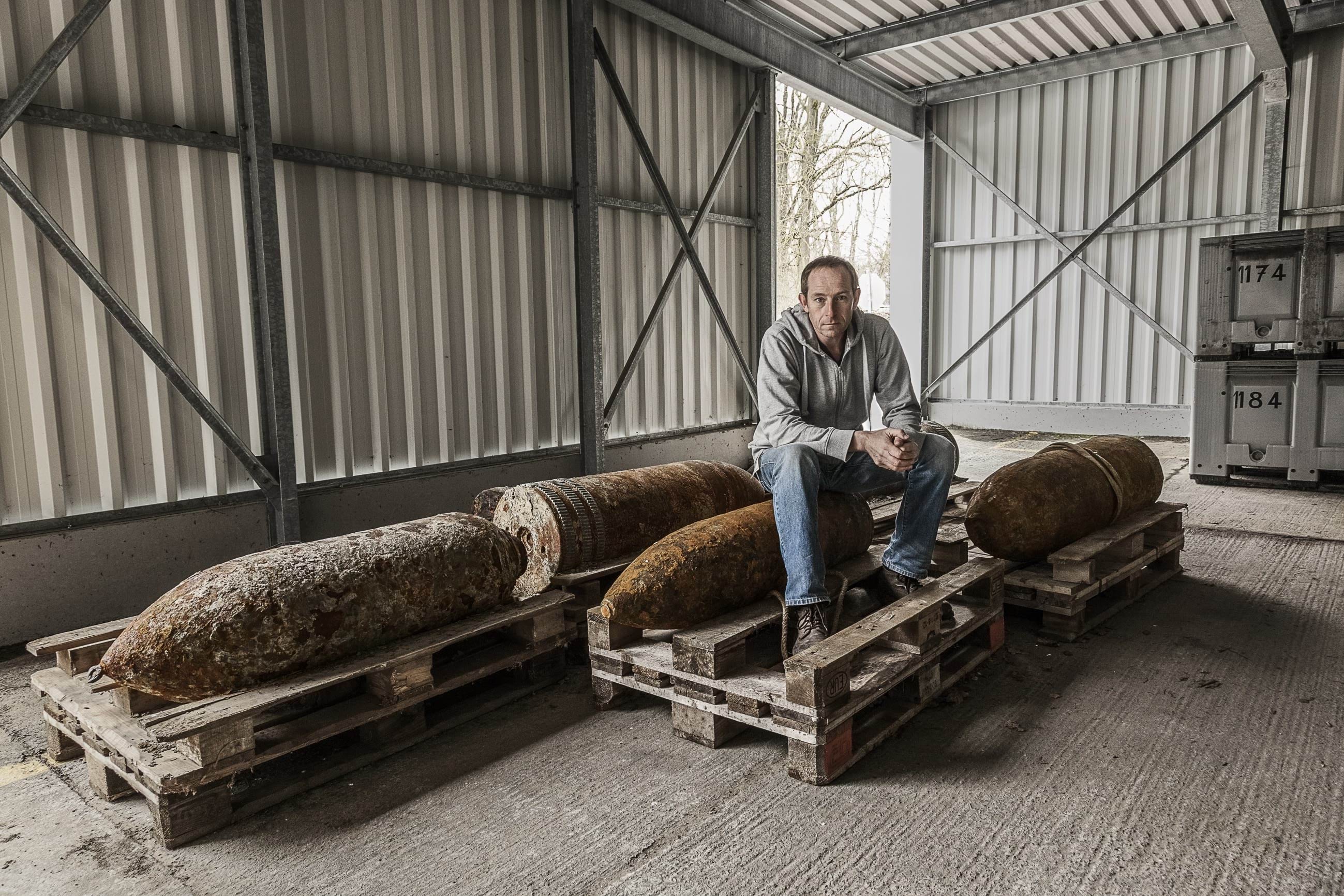 A man in a grey jumper and blue jeans sitting on recovered ammunition from the First World War. He is staring straight into the camera with a solemn look on his face. Around him are three other large shells on wooden crates.