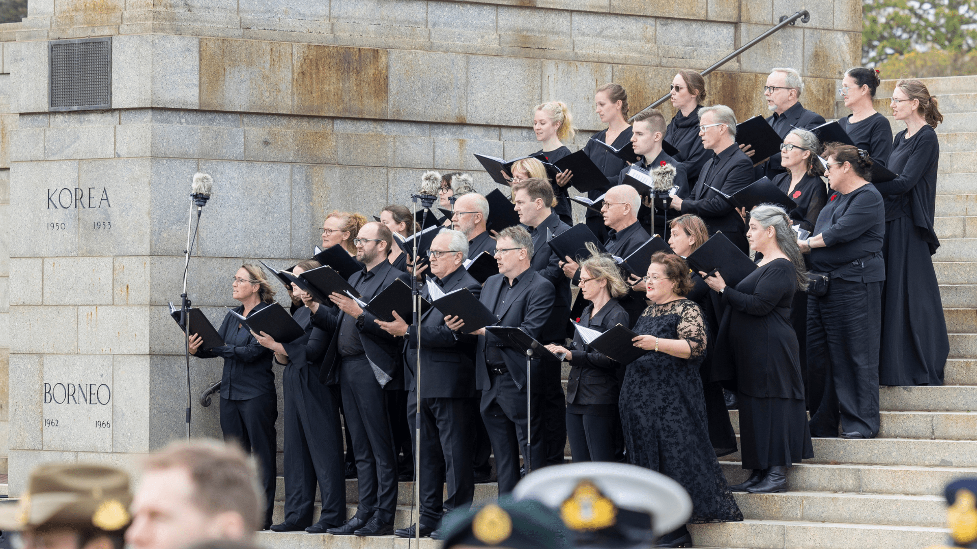 A choir standing on the steps of the Shrine of Remembrance. They are all wearing black with red poppies on their lapel. They are all holding songbooks and singing toward their conductor. 